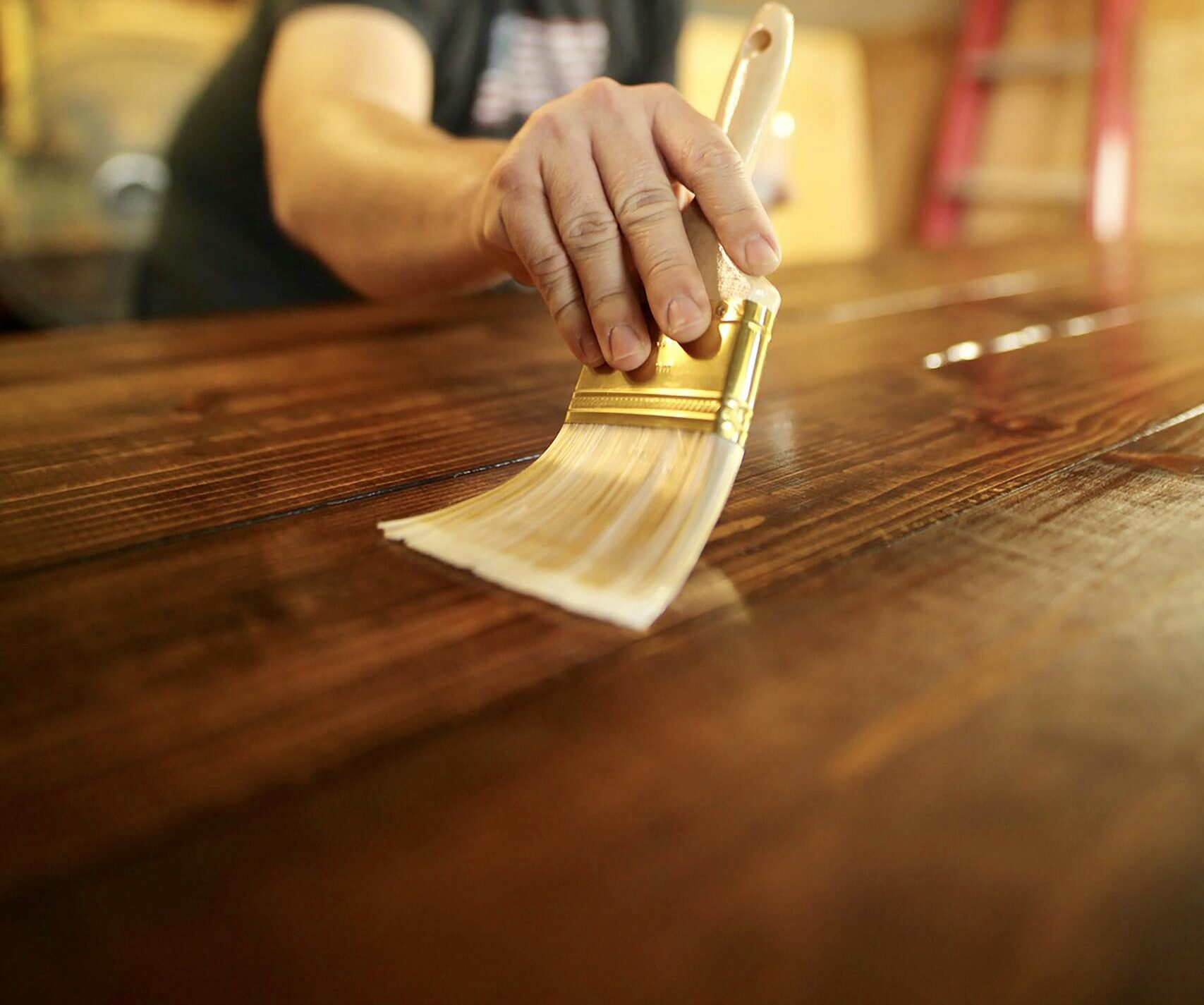 A man carefully applies varnish to a wooden surface indoors, using a paintbrush for a smooth finish.